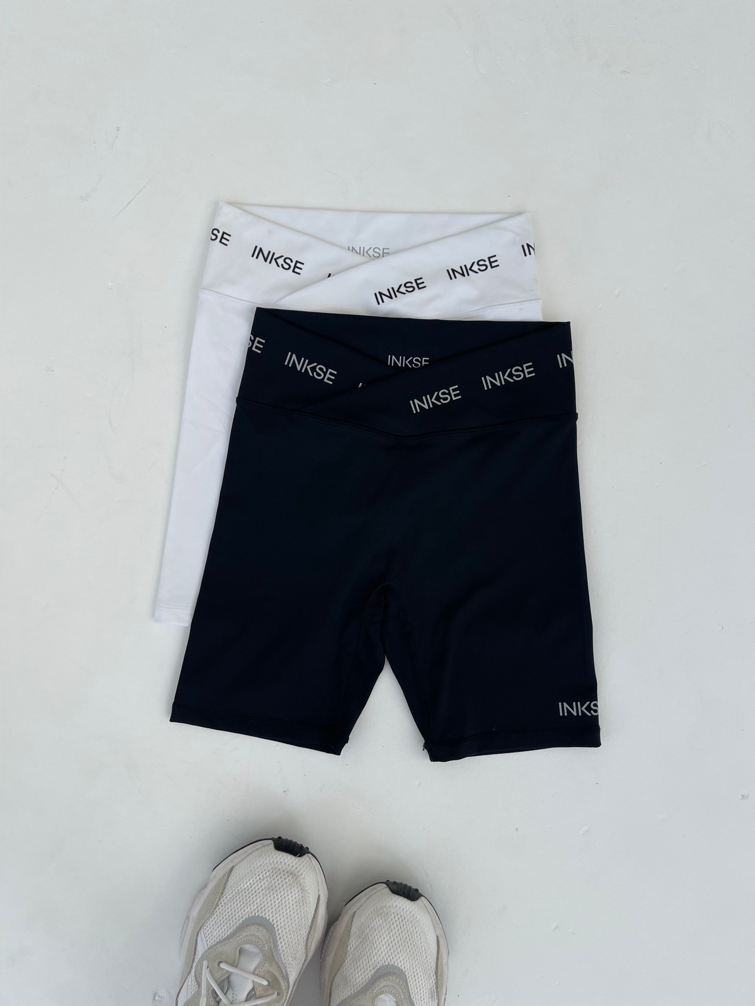 Black and white custom bike shorts with a branded logo waistband laid flat beside sneakers